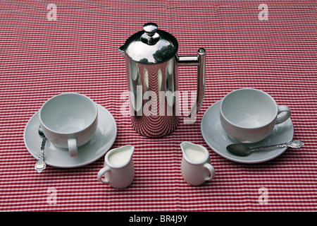 A shiny coffee pot and cups and saucers on a red and white tablecloth Foto Stock