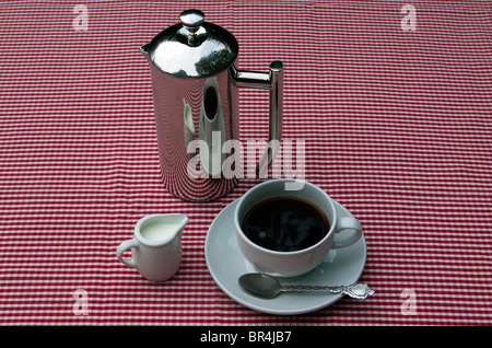 A shiny coffee pot and cups and saucers on a red and white tablecloth Foto Stock