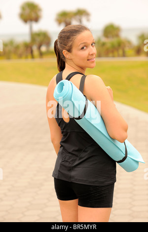 Bellissimo modello fitness portando materassino yoga in una panchina nel parco vicino alla spiaggia. Foto Stock