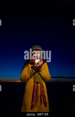 Un sorridente giovane donna detiene una tazza di cioccolata calda nella parte anteriore di un Cielo di tramonto Foto Stock