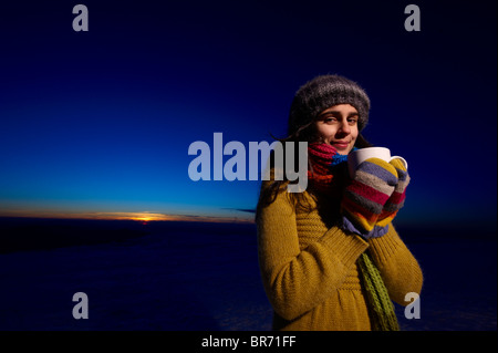 Un sorridente giovane donna detiene una tazza di cioccolata calda nella parte anteriore di un Cielo di tramonto Foto Stock