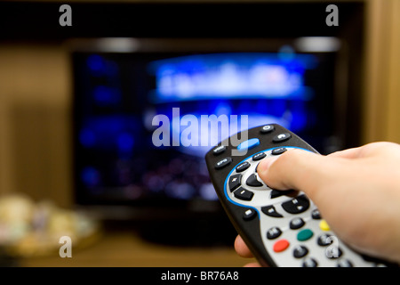 Mano che tiene il telecomando del TV con una televisione in background. Close up. Foto Stock