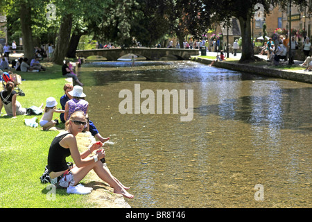 Gente seduta sul greto del fiume a Bourton-on-the-acqua nel Gloucestershire conosciuta come la "Venezia del Costwolds' Foto Stock