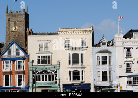 Grazioso negozio colorata fronti e il campanile di una chiesa sulla banchina a Dartmouth, Devon, con un cielo blu e St George battenti bandiera Foto Stock