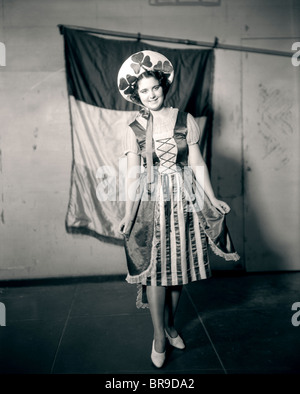 1910s 1920 giovane donna indossa l'Irlanda COSTUME NAZIONALE CON TRIFOGLI SU HAT guardando la telecamera bandiera irlandese IN BACKGROUND Foto Stock