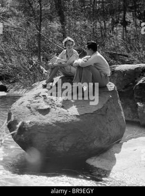 1930s giovane uomo donna seduta sulla grande roccia nel flusso abbracciando le ginocchia sorridendo felice romanticismo Foto Stock