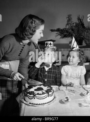 Anni Cinquanta donna madre il taglio di torta di compleanno per due bambini seduti a tavola indossando cappelli di partito Foto Stock