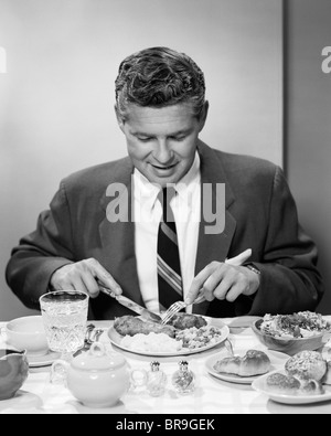 Anni Cinquanta uomo sorridente in tuta e cravatta seduta a tavola tenendo il coltello e la forchetta di mangiare la cena Foto Stock