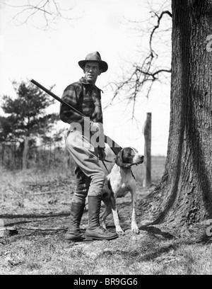 1930s uomo HUNTER INDOSSANDO HAT PLAID FLANNEL SHIRT I JODHPURS e stivali in piedi tenendo un fucile con cane da ferma Foto Stock