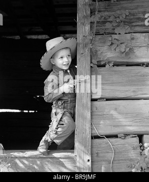 Anni Cinquanta BOY IN WESTERN cowboy abito cappuccio di tenuta pistola a giocare nel vecchio portone in legno Foto Stock