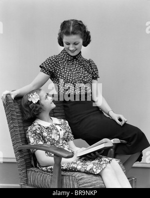 1920s 1930 sorridente bambina guardando alla sorella donna seduta su una sedia la lettura Foto Stock