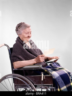 1960 anni sessanta retrò senior sorridente anziani donna seduta in carrozzella LETTURA LA LETTERA DI ASSICURAZIONE SANITARIA VERIFICARE Foto Stock