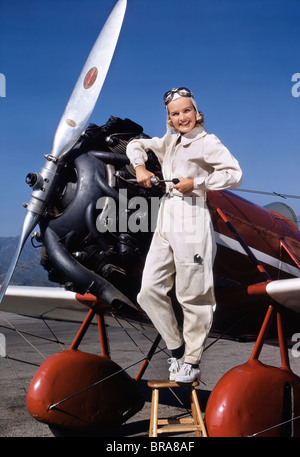 1930s 1940s donna sorridente aereo MECCANICO PILOTA IN TUTE STAND LAVORANDO SUL MOTORE elica guardando la fotocamera Foto Stock