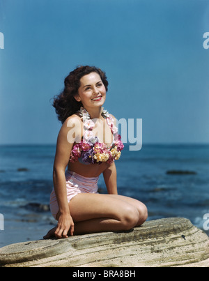 Negli anni quaranta anni cinquanta sorridente BRUNETTE donna seduta su roccia che indossa due pezzi COSTUME DA BAGNO APPLIQUÉD con fiori di colore pastello Foto Stock