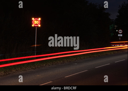 Lampeggiante 30mph segno di velocità in un villaggio di auto e le luci di coda di notte. Esposizione lunga Foto Stock