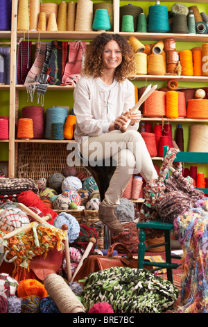 Donna seduta su uno sgabello Holding degli aghi di tessitura nella parte anteriore del display del filato Foto Stock