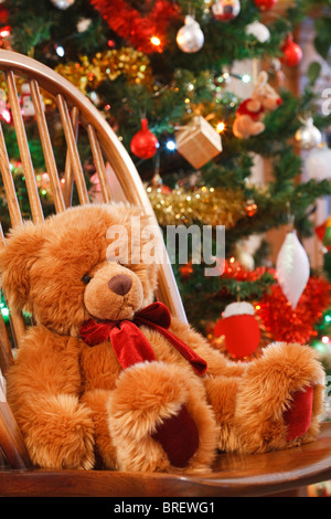 Natale interno con un orsacchiotto di peluche su una sedia davanti a un albero di natale Foto Stock