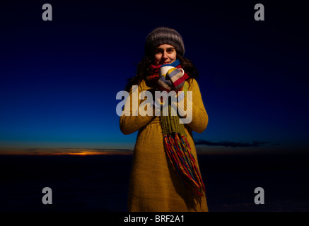 Un sorridente giovane donna detiene una tazza di cioccolata calda nella parte anteriore di un Cielo di tramonto Foto Stock