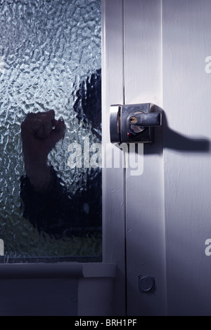 Silhouette di un intruso maschio di guardare attraverso una finestra opaca accanto a una porta anteriore Foto Stock