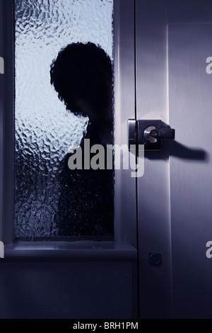 Silhouette di un intruso maschio di guardare attraverso una finestra opaca accanto a una porta anteriore Foto Stock