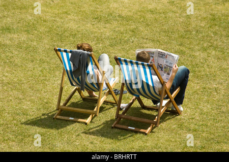 Antenna orizzontale vista posteriore di un giovane uomo e di una donna, rilassarsi e prendere il sole in un parco seduto su striped sdraio in legno Foto Stock