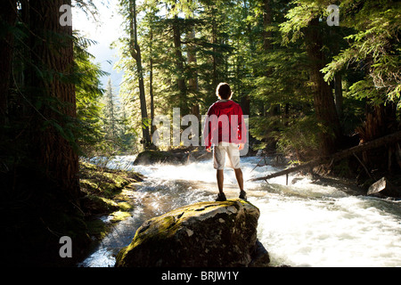 Un giovane uomo di escursionismo sorge su una roccia e orologi il flusso di fiume della foresta. Foto Stock