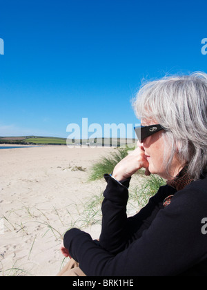 Donna anziana seduta sulla spiaggia di Rock, Cornovaglia, Inghilterra. Foto Stock