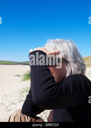 Donna anziana seduta sulla spiaggia di Rock, Cornovaglia, Inghilterra. Foto Stock