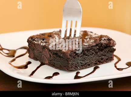 Uno ricco di spessore brownie al cioccolato con una forcella inserita su una piastra rivestita in sciroppo spruzzata con uno sfondo giallo Foto Stock