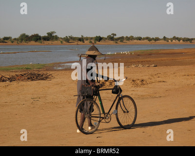 L'uomo africano con il suo ritorno alla telecamera, indossando un cappello tradizionale spingendo una bicicletta lungo una riva di un fiume Foto Stock