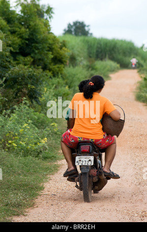 Donna in T-shirt riding pillion su una vecchia moto giù per una strada sterrata in Isaan, nel nordest della Thailandia. Foto Stock