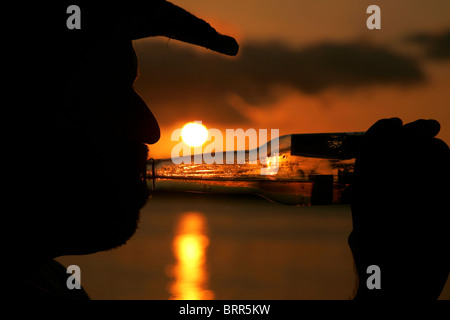 Vista laterale la silhouette di un uomo di bere da una bottiglia di vetro con una impostazione sun in background Foto Stock