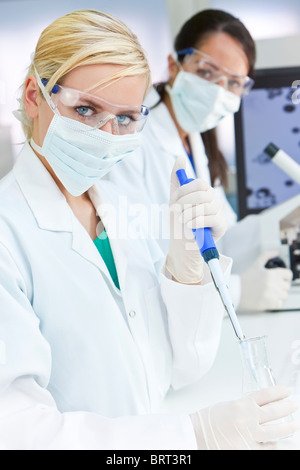 Una femmina di medici o di ricerca scientifica o scienziato usando una pipetta e il pallone in un laboratorio con il suo collega dietro di lei Foto Stock
