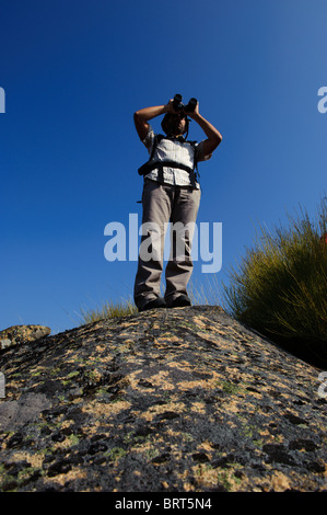 Giovane uomo con zaino guardando attraverso il binocolo sulla sommità di una roccia contro il cielo Foto Stock