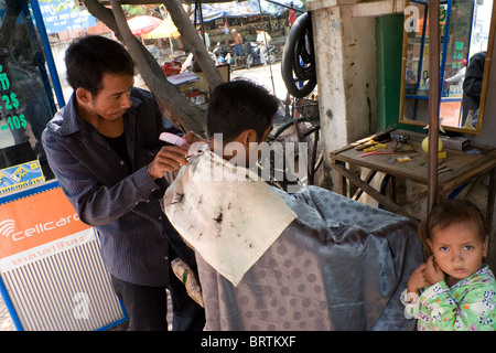 Un barbiere di strada è il taglio di un uomo di capelli come una giovane ragazza è in piedi vicino a una strada barbiere in Phnom Penh Cambogia. Foto Stock