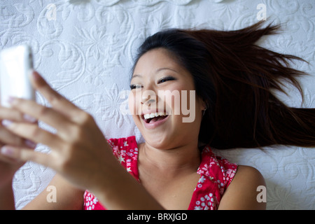 Sorridente razza mista ragazza adolescente posa sul letto e di messaggistica di testo sul telefono cellulare Foto Stock