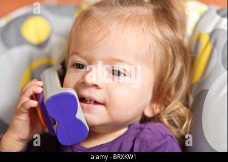 Un bambino ragazza seduto in una sedia alta finge di essere in possesso di una conversazione su un giocattolo mobile telefono. Foto Stock