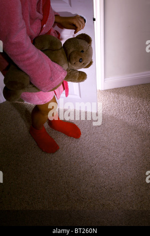 Bambina che impugnano un orsacchiotto di peluche in piedi dietro una porta aperta. Modello e proprietà (proprietà di fotografo) rilasciato. Foto Stock
