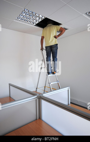 Uomo sulla parte superiore della scaletta di valutare un problema elettrico nel controsoffitto di un ufficio Foto Stock