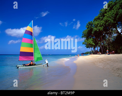 Uomo che lancia il catamarano colorato a vela dal bordo delle acque della spiaggia di sabbia tropicale con coppia di vacanza a bordo a Tamarind Cove Barbados Caraibi Foto Stock