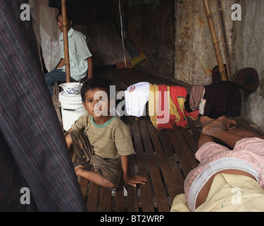 Un giovane bambino operaio ragazzo che vivono in condizioni di povertà è seduta su di un letto in legno mentre un altro ragazzo è dormire in Kampong Cham, Cambogia. Foto Stock