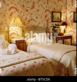 Lampade illuminate su entrambi i lati di due letti singoli con piumino floreale in camera da letto tradizionale con rosso + bianco carta da parati Foto Stock