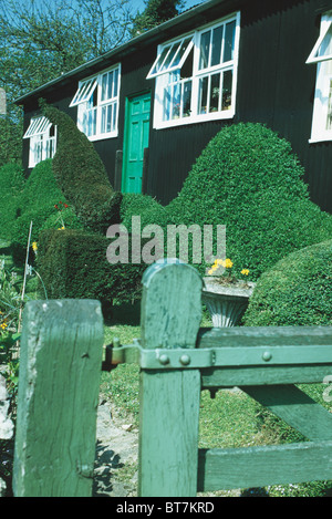 Il verde cancello di legno per giardino con topiaria da arbusti di fronte ad un piano singolo nero casa in legno con finestre bianche Foto Stock