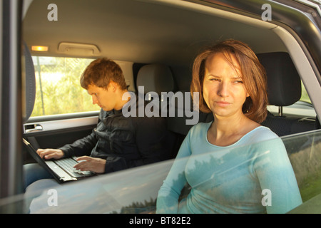 La donna guarda attentamente fuori dal finestrino dell'auto mentre l'uomo accanto a lei lavora sul laptop sul sedile posteriore Foto Stock