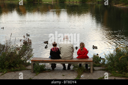 Nonno e i suoi due nipoti a sedersi su una panchina presso il lago di anatre di alimentazione Foto Stock