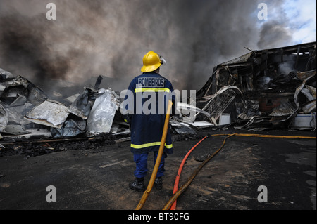 Lone fireman sembra impotente a resti di un edificio di fabbrica bruciata da un massiccio fuoco Foto Stock