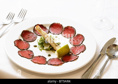 Fette di carne affumicata su una piastra bianca con argenti e bicchiere di vino. Orientamento orizzontale, pulito, semplice, sfondo bianco. Foto Stock