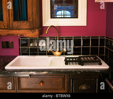 Close-up di mattonelle nere splash-torna al di sopra di ottone rubinetto miscelatore e bianco lavello da cucina in legno montati unità in cucina rosa Foto Stock