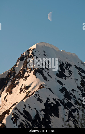 Una mezza luna passa sopra una neve-crested picco nella valle di Portage, Chugach National Forest, centromeridionale Alaska, molla Foto Stock