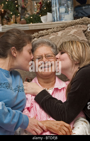 Due nipoti adolescenti baciare la loro nonna sulle sue guancie mentre si è seduti sul divano con decorazioni di Natale in background. Foto Stock
